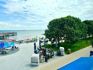A waterfront café with outdoor seating, a pier in the background, and a large tree providing shade. - House With sea View - Apt. 3 (Wyk auf Foehr)