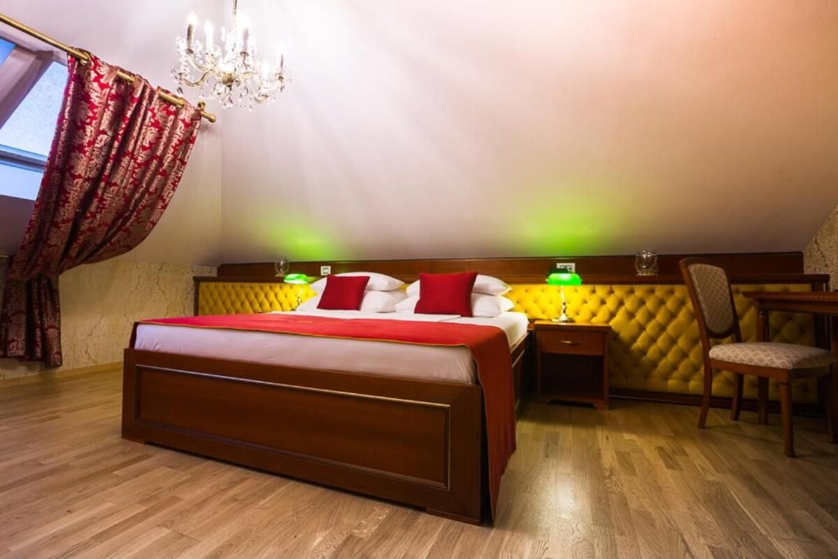 Ein Hotelzimmer mit einem großen Bett, einem Nachttisch, einem Sessel, einer Chandelüster und einem Fenster mit Vorhängen.