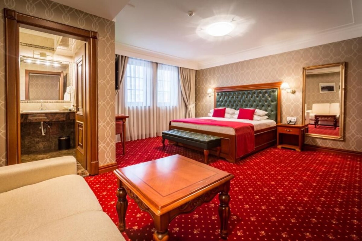 Ein Hotelzimmer mit einem großen Bett, einem Schreibtisch, einer Bank, einem Spiegel und einem Badezimmer mit Waschbecken und Spiegel.