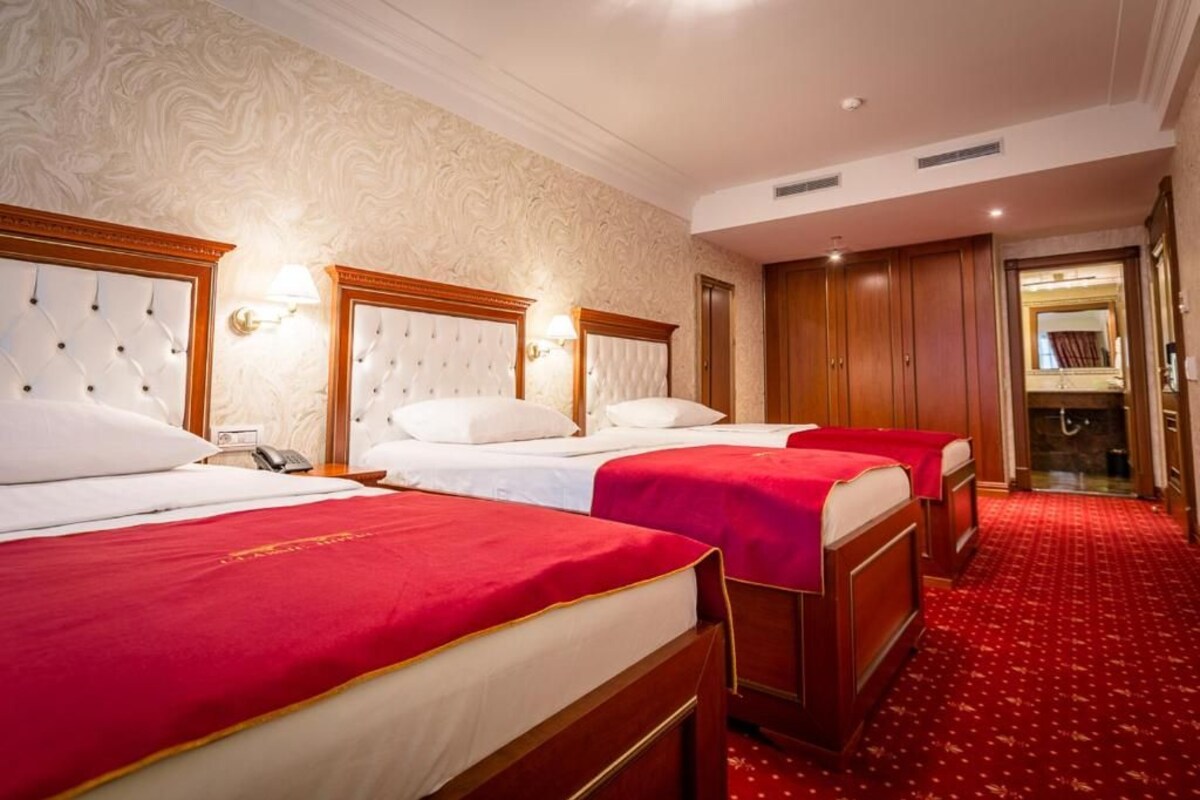 Ein Hotelzimmer mit drei Betten, jeweils mit roter Bettdecke und weißen Kissen, und einem hölzernen Kopfteil. Auf dem Nachttisch steht ein Telefon und an der Wand ist eine Lampe angebracht.