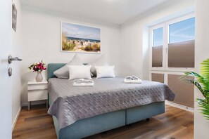A bedroom with a bed, pillows, a nightstand with a vase of flowers, a window with blinds, and a painting on the wall. - Beach Runner 4.2 | Sauna. Patio. Near the beach. (Kellenhusen)
