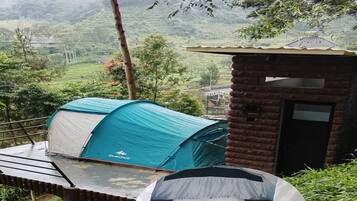 A tent with a sleeping area and a small brick structure, set against a hilly landscape.