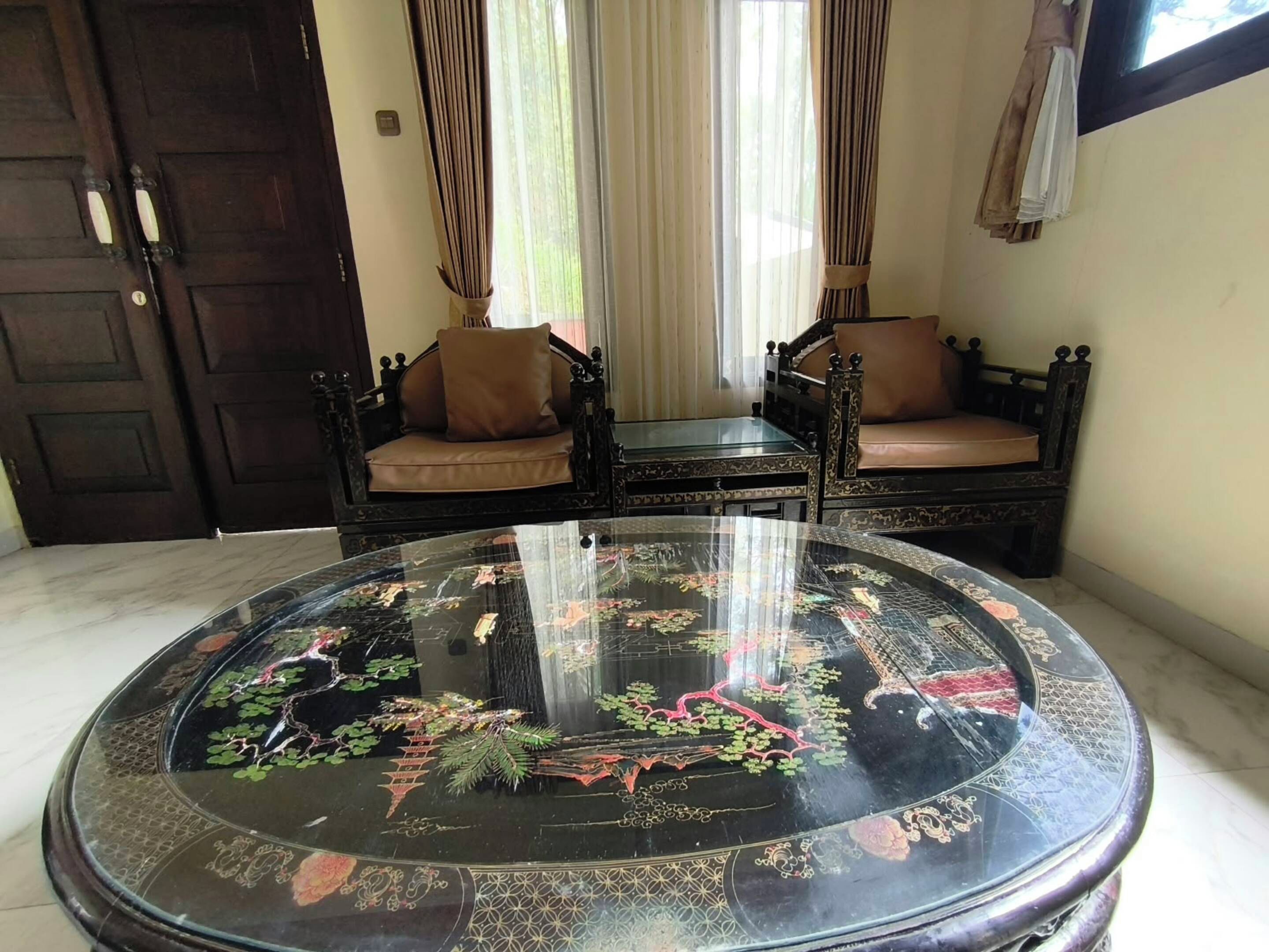 A room with a round glass-top table featuring intricate designs, two dark wooden sofas with cushions, a closed dark wooden door, and a window with sheer curtains.