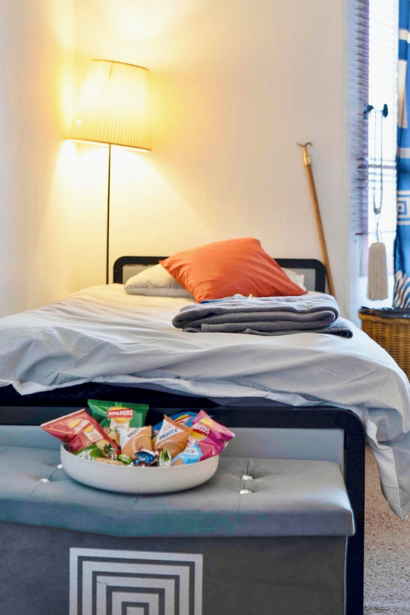 Una cama bien hecha con un almohadón, una mesita de noche con aperitivos y una lámpara en una habitación con ventana.
