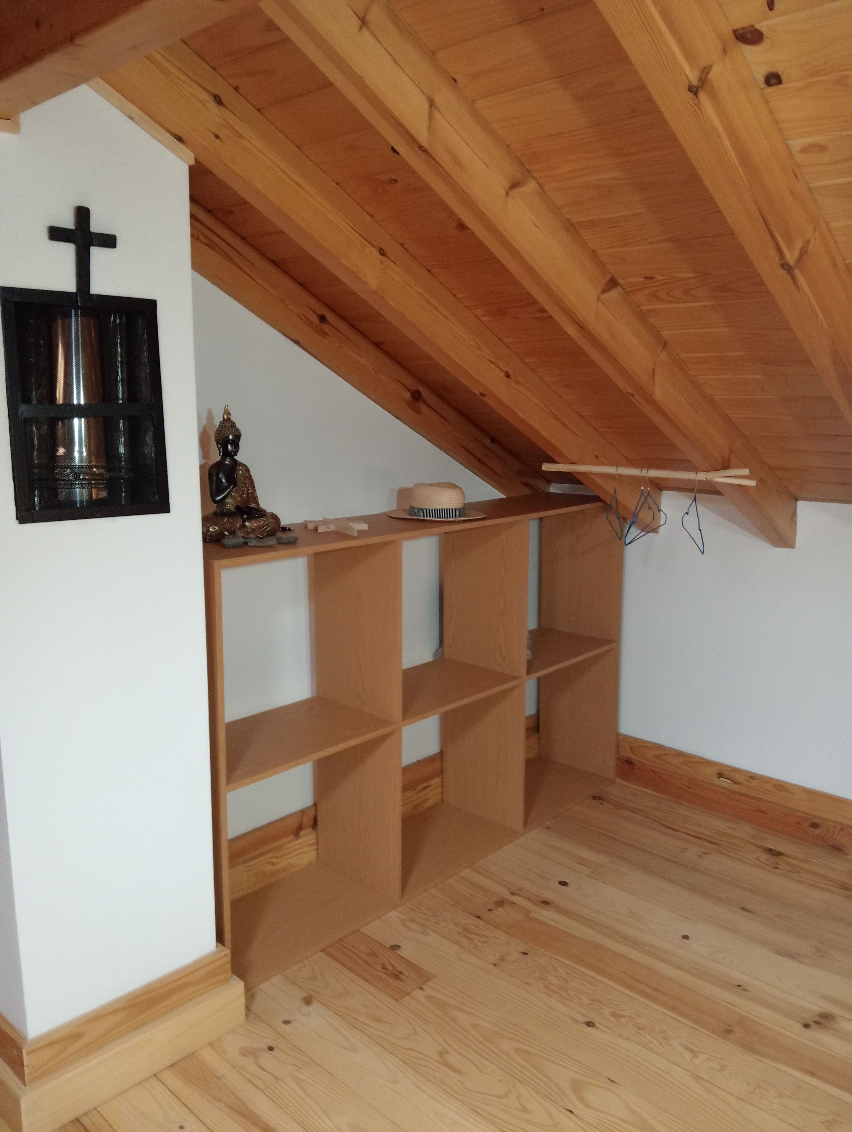A room with a sloped ceiling, wooden beams, a cross on the wall, and a shelf with a Buddha statue and a hat.