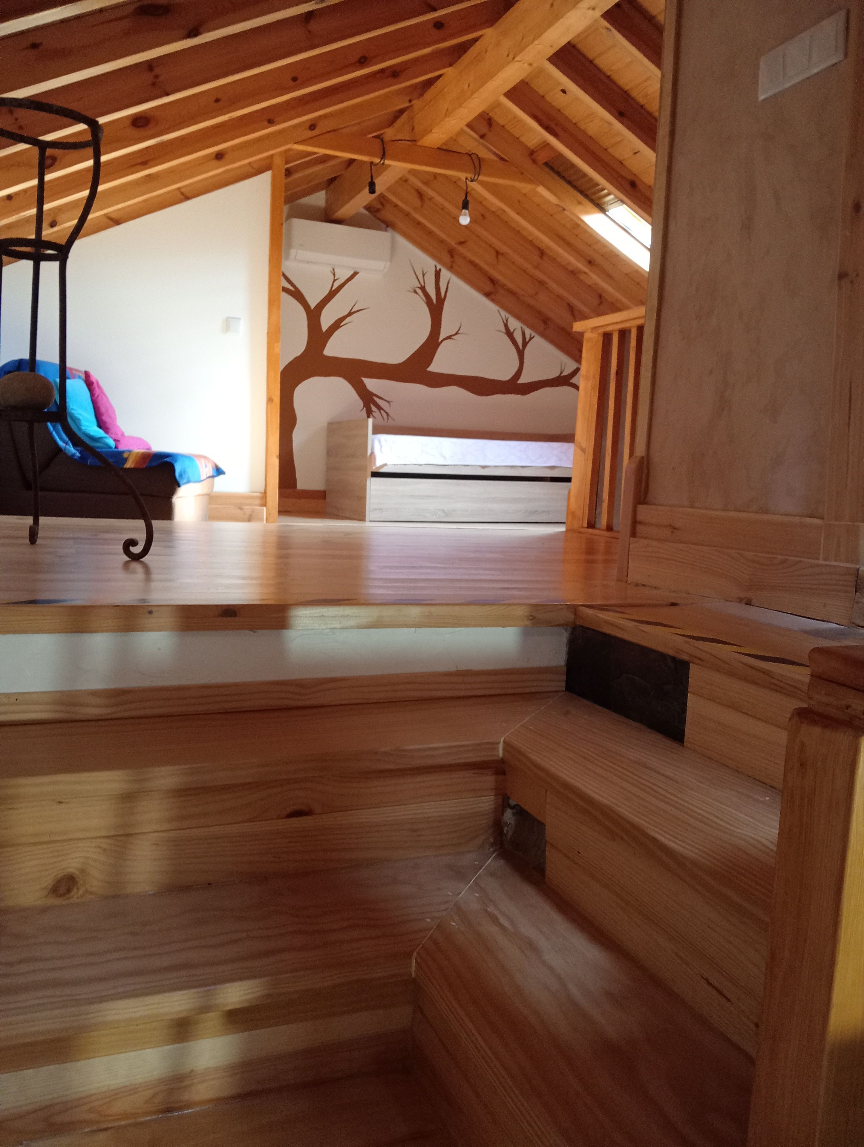 A room with a sloped ceiling, wooden beams, a bed, a chair, and a wall mural.