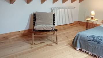 Ein Zimmer mit schrägen Wänden, einem Holzstuhl, einem Bett mit Decke, einem Heizkörper und einem kleinen Tisch mit Lampe.