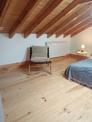 A room with a sloped ceiling, a wooden chair, a bed with a blanket, a radiator, and a small table with a lamp.