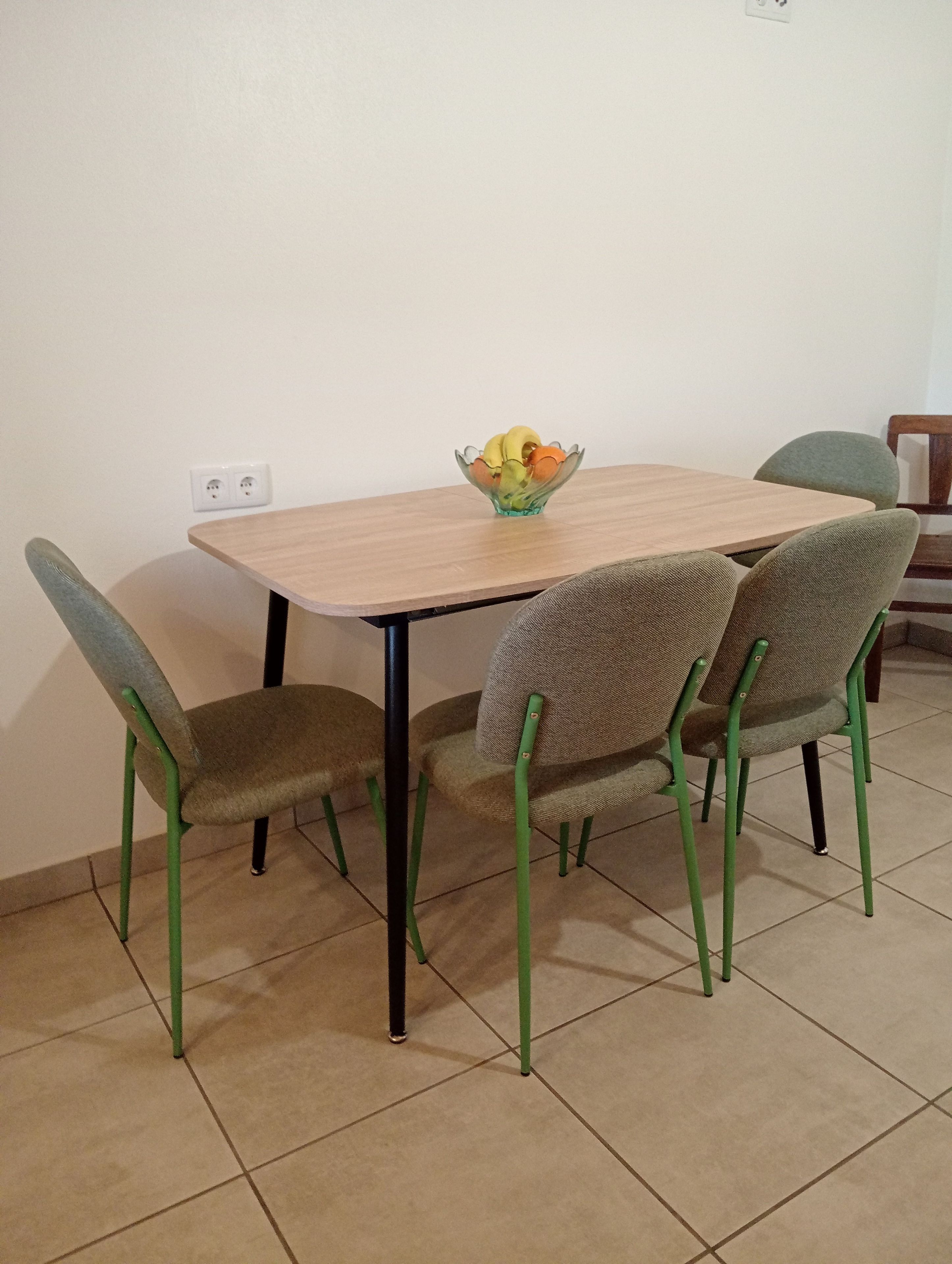 A dining table with four chairs, a bowl of fruit, and a power outlet on the wall.