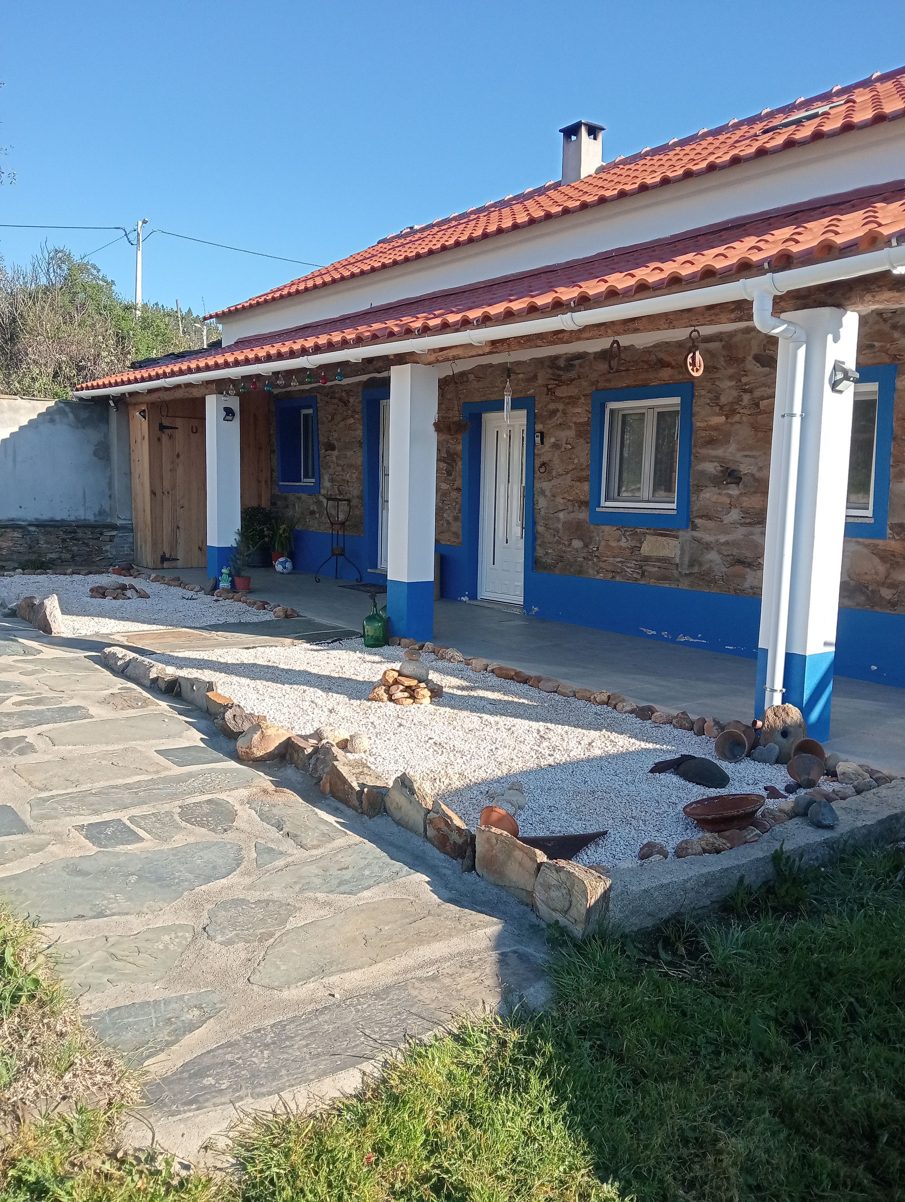 A two-story building with a red-tiled roof, stone walls, and blue trim. There is a paved area with rocks and a small garden.