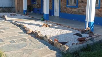 A two-story building with a red-tiled roof, stone walls, and blue trim. There is a paved area with rocks and a small garden.