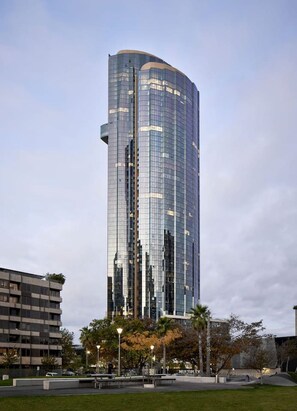 A tall, modern glass building with a curved facade, surrounded by a landscaped area with trees and a paved walkway. - Stunning Southbank 2 Bedroom (Southbank)