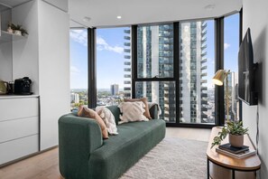 A modern living room with a green sofa, a coffee machine, a TV, and a view of high-rise buildings. - Stunning Southbank 2 Bedroom (Southbank)