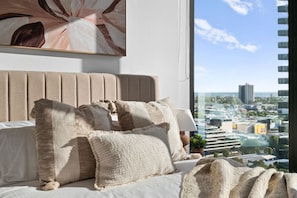 Ein Schlafzimmer mit großem Fenster, einem Bett mit beige farbenem Bettzeug und Blick auf die Stadt.