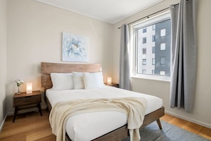 A neatly made bed with a wooden headboard, a bedside table with a lamp, and a window with curtains. - Central Apartment with Tennis (Southbank)