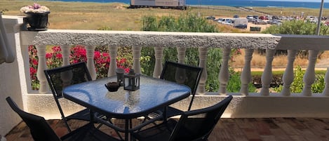 A glass-top table with four chairs on a balcony overlooking a beach and a parking lot.