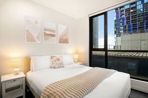 A hotel room with a large bed, two bedside tables, a window with a city view, and three wall-mounted abstract paintings. - Light-filled City 2 Bedroom (Melbourne)