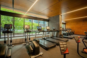 A gym with treadmills, elliptical machines, and stationary bikes, large windows with a view of greenery, and a modern design with wooden paneling. - Designer Urban 2 Bedroom (Melbourne)