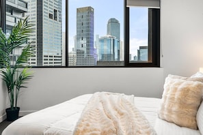 A bedroom with a large window, a bed with white bedding, a fluffy throw, and a potted plant. - Designer Urban 2 Bedroom (Melbourne)