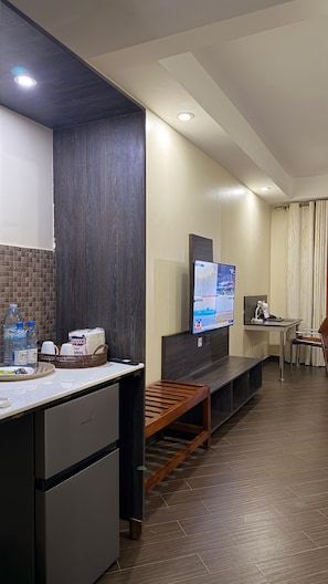 A modern hotel room with a kitchenette, a flat-screen TV mounted on the wall, a wooden bench, and a small table with a chair. - NEXUS RESORTS HOTEL (Kampala)