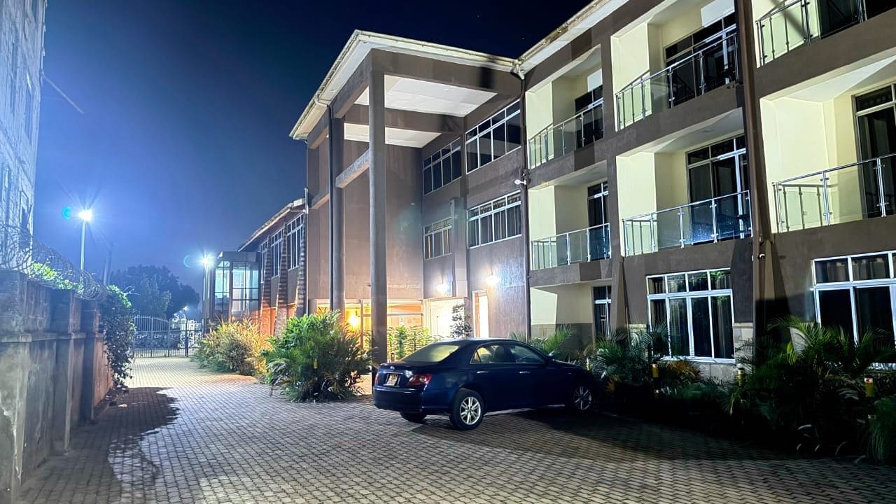 A multi-story building with a car parked in front, surrounded by greenery and a paved walkway.