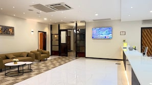 A modern hotel lobby with a reception desk, a sofa, a coffee table, and a TV screen displaying a news broadcast. - NEXUS RESORTS HOTEL (Kampala)