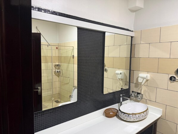 Un baño con ducha envuelta en vidrio, un lavamanos blanco con un recipiente decorativo, y un espejo sobre el lavamanos.