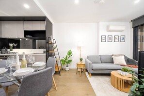 A modern living room with a grey sofa, a dining table set with plates and glasses, a coffee table, and a small side table with a lamp. - Super Central 2 Bedroom (Melbourne)