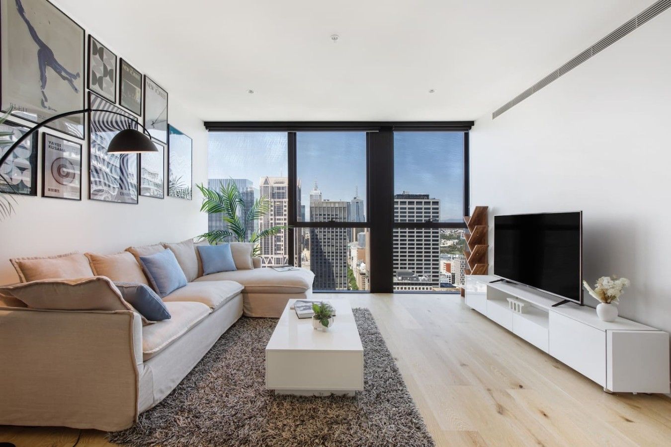 A modern living room with a sectional sofa, a flat-screen TV, and a large window offering a city view.