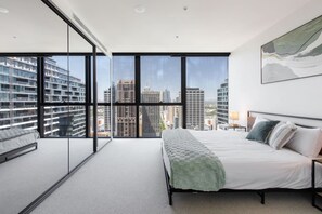 A modern bedroom with a large bed, a view of the cityscape through floor-to-ceiling windows, and a minimalist design. - New York Style City 3 Bedroom (Melbourne)