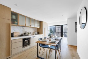 A modern kitchen with wooden cabinets, a dining table with chairs, and a large window offering a city view. - New York Style City 3 Bedroom (Melbourne)