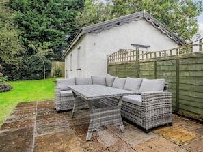 A patio with a wicker sofa and table, a stone floor, and a white building with a green fence in the background. - Waltergarth (Settle)