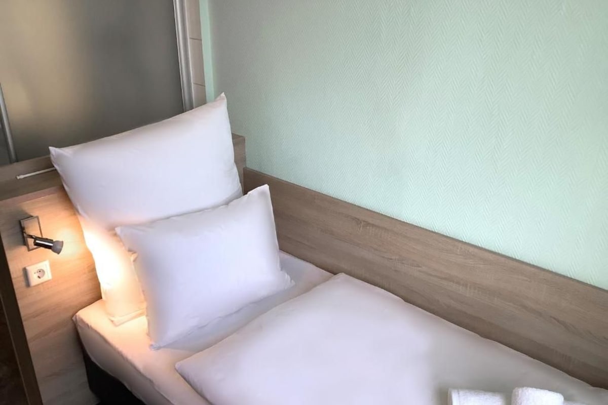 Een net opgemaakt bed met witte lakens en kussens, een bedlampje en een handdoek op de vloer.