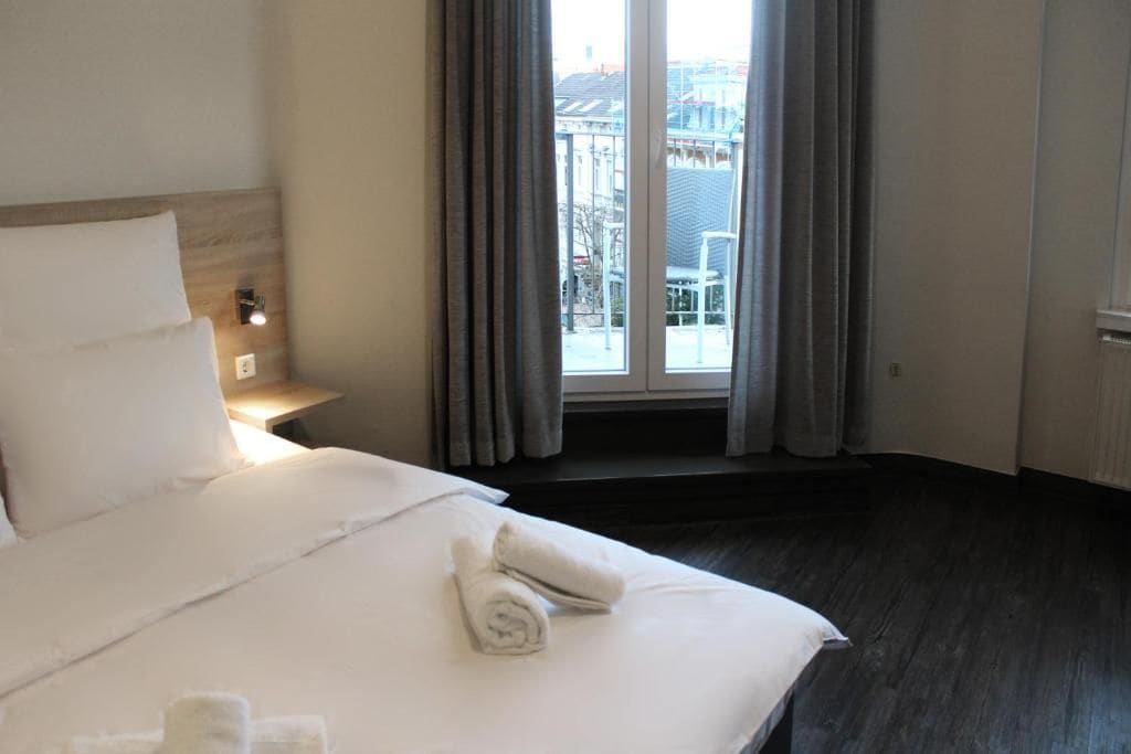 A hotel room with a large bed, a window with curtains, a nightstand with a lamp, and a view of buildings outside.