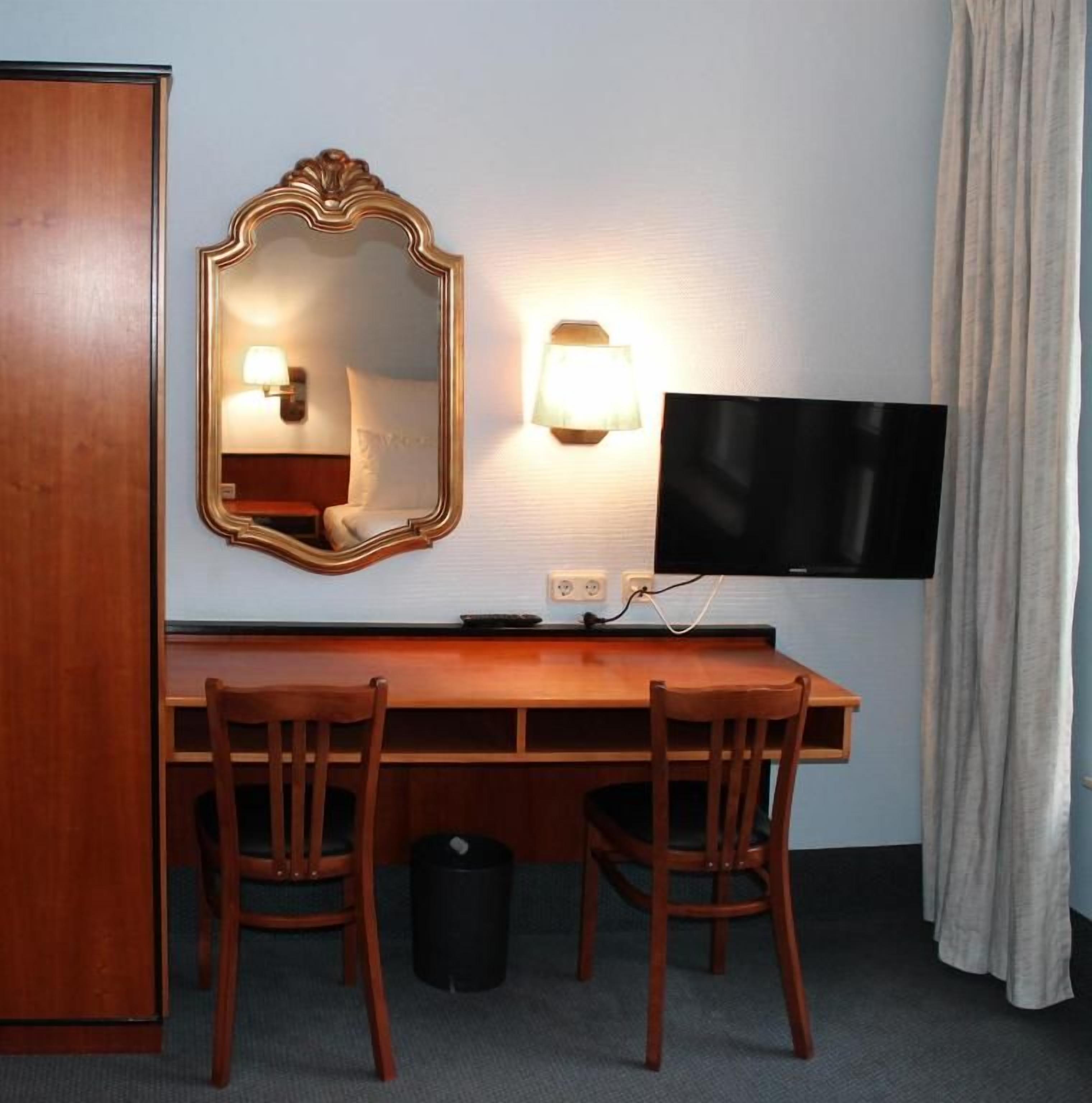 A hotel room with a wooden desk, two chairs, a television, a wall-mounted mirror, and a lamp.