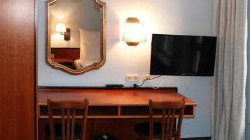 A hotel room with a wooden desk, two chairs, a television, a wall-mounted mirror, and a lamp.