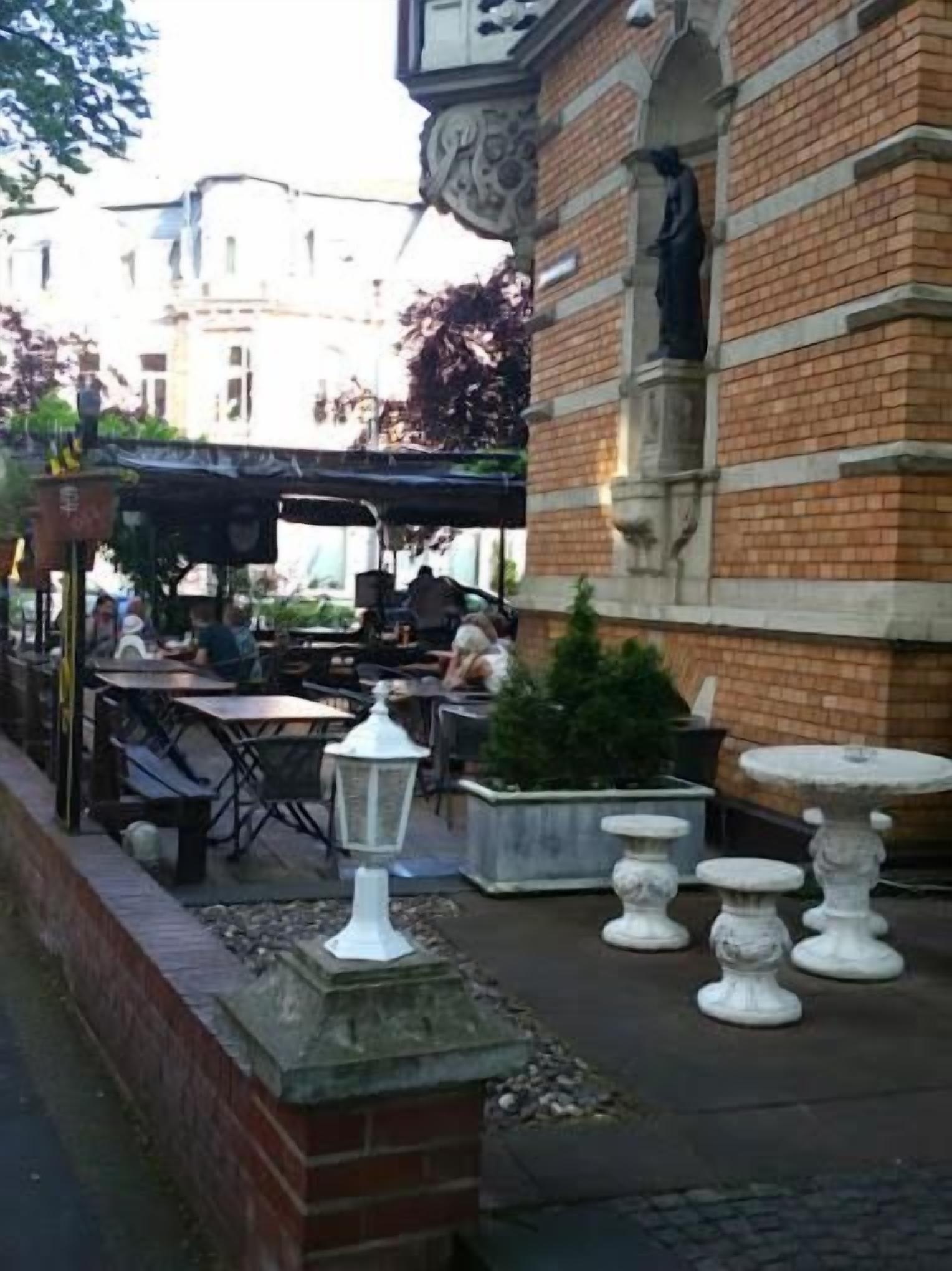 An outdoor seating area with tables and chairs, a white lantern, and a brick building with a statue.