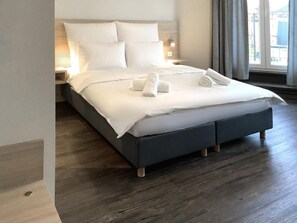 A neatly made bed with white linens and towels, a wooden floor, and a nightstand with a lamp.