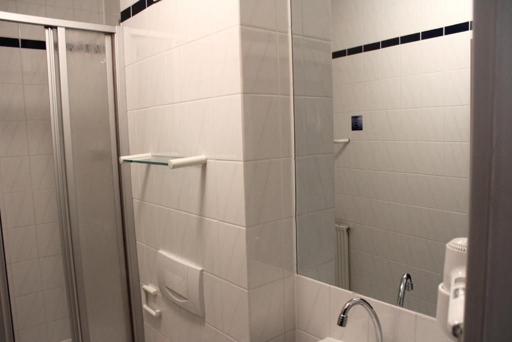 Ein Badezimmer mit weißen Fliesen, einer Duschkabine aus Glas, einem Spiegel und einem Waschbecken mit Wasserhahn.