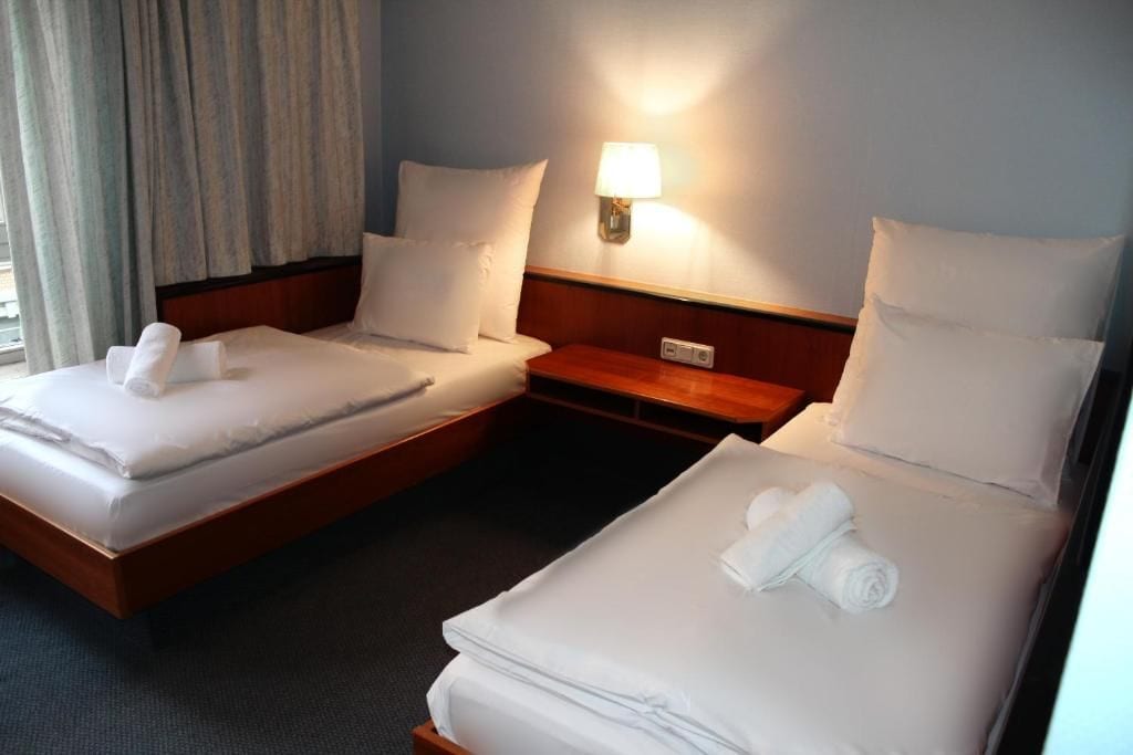 Ein Hotelzimmer mit zwei Betten, jeweils mit weißer Bettwäsche und Kissen, einem Nachttisch mit Lampe und einem Vorhangfenster.