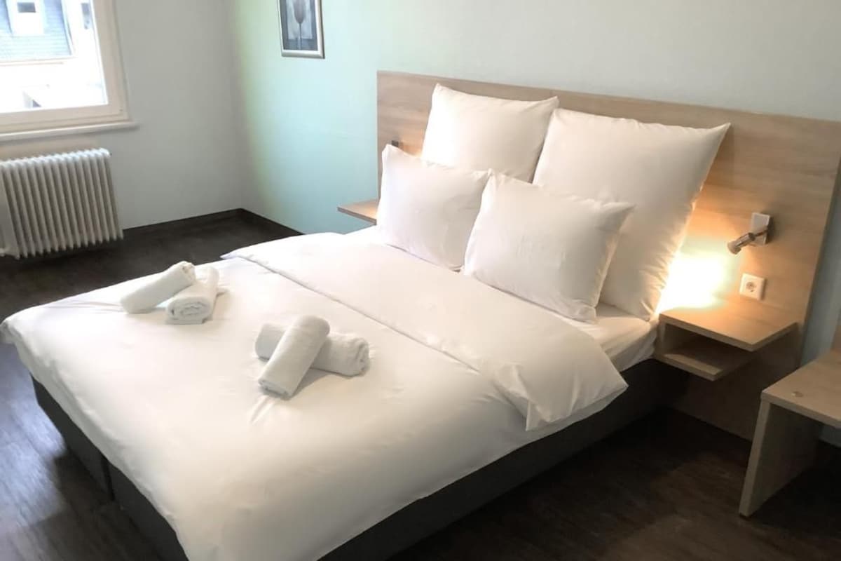 Een netjes opgemaakt bed met witte lakens en kussens, een nachtkastje met een lamp, en een radiator op de achtergrond.