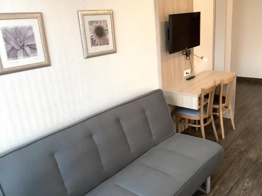 A room with a grey sofa, a wooden dining table with chairs, a flat-screen TV mounted on the wall, and framed pictures on the wall.