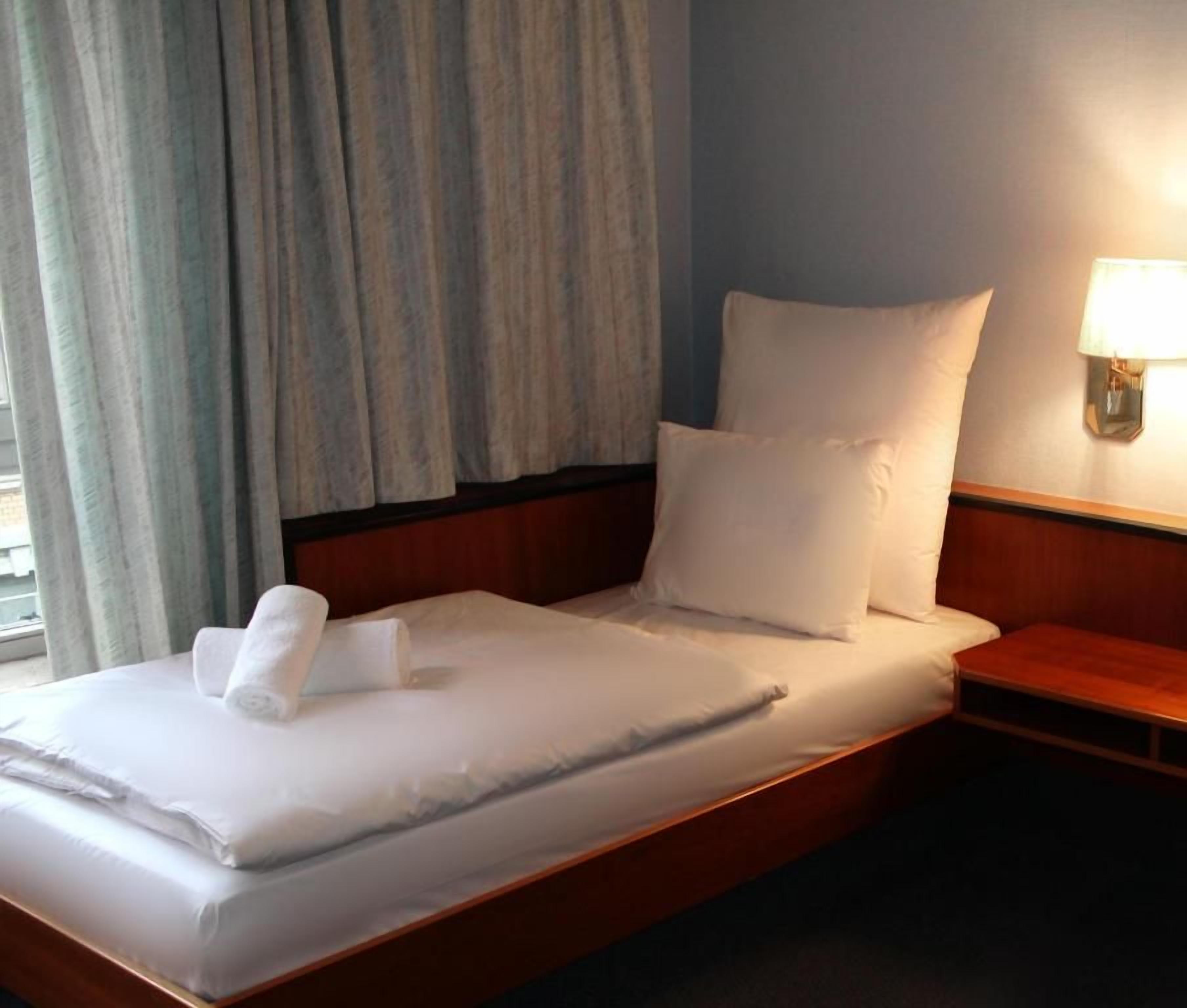 Habitación de hotel con una cama, una mesita de noche, una lámpara y un ventanal con cortinas.