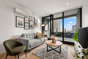 A modern living room with a grey sofa, a round coffee table, and a large window offering a city view. - Elegant 2 Bedroom with Views (Docklands)