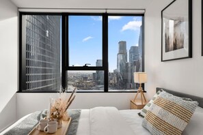 Een slaapkamer met een groot raam, een bed, een nachtkastje met een lamp en een lijst met een afbeelding aan de muur.