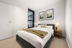 A modern bedroom with a large bed, a window, two framed pictures, a bedside table with a lamp, and a door. - Elegant 2 Bedroom with Views (Docklands)