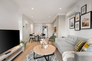 A modern living room with a flat-screen TV, a grey sofa, a round coffee table, and a dining area with chairs and a table. - Elegant 2 Bedroom with Views (Docklands)
