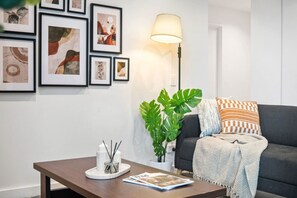 A living room with a dark sofa, a coffee table with a magazine, a floor lamp, and framed artwork on the wall. - Urban Treehouse Oasis (Docklands)