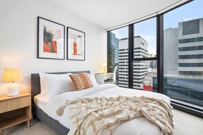 A modern hotel room with a large bed, a nightstand, a desk, and a view of the cityscape through large windows. - Urban Treehouse Oasis (Docklands)