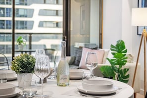 Une table à manger dressée avec des assiettes, des verres et une bouteille, avec en arrière-plan une vue sur un bâtiment et une plante.